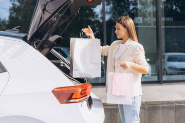 Başarılı bir alışveriş gezisinden sonra alışveriş çantalarını bagajına dolduran şık genç bir kadın. Güneşli bir günde beyaz bir arabanın yanında duruyor, mutluluk ve memnuniyet yayıyor..