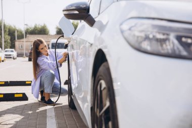 Bir kadın elektrikli aracını güneşli bir kentsel bölgedeki şarj istasyonuna bağlıyor, sürdürülebilir ulaşım ve çevre dostu çözümleri vurguluyor. Bu sahne, modern çevre bilincinin özünü yakalar..