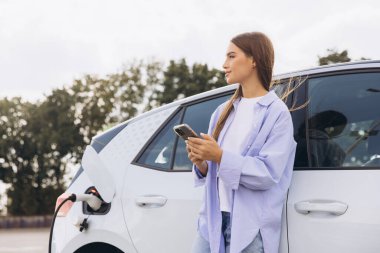 Genç bir kadın elektrikli arabasının yanında duruyor, akıllı telefonunu kullanıyor. Araba şarj oluyor, çevre dostu ve modern teknolojiyi sembolize ediyor. Sahne, bağlantı ve sürdürülebilir yaşamı tasvir ediyor.