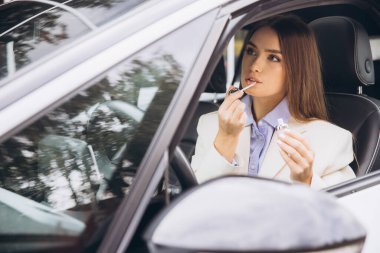 Arabada oturan şık bir kadın, dudak parlatıcısını titizlikle sürüyor. Kendine güveni ve entellektüelliği var. Beyaz ceket ve mavi gömlek giyip bir anlık zarafeti yakalıyor..