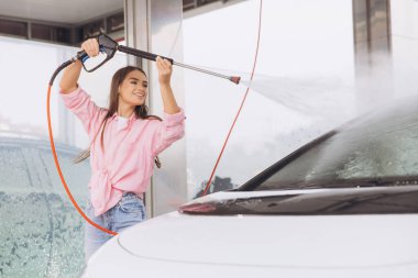 Neşeli genç bir kadın otomobil yıkama istasyonunda elektrikli çamaşır makinesiyle arabasını mutlulukla yıkıyor. Kurulum, bağımsızlık, temizlik ve araç bakımıyla tatmin duygusunu teşvik eder.