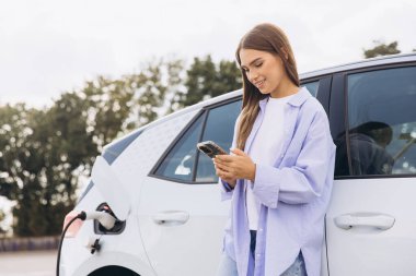 Genç bir kadın elektrikli arabasının yanında duruyor, akıllı telefonunu kullanıyor. Araba şarj oluyor, çevre dostu ve modern teknolojiyi sembolize ediyor. Sahne, bağlantı ve sürdürülebilir yaşamı tasvir ediyor.