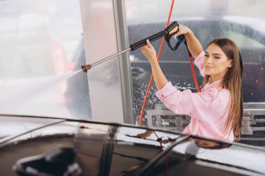 Genç bir kadın self-servis istasyonunda arabasını yıkamaktan hoşlanır. Gülümsüyor, yüksek basınçlı su hortumu kullanıyor ve pembe bir gömlek giyiyor..