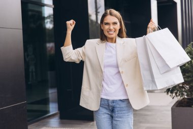 Neşeli kadın dışarıda elinde alışveriş torbalarıyla kutlama yapıyor. Başarılı bir alışveriş çılgınlığından sonra mutluluk ve güven yayar. Perakende, moda veya yaşam tarzı temaları için mükemmel görüntü.