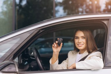 Kendine güvenen bir kadın, modern aracının içinde arabanın anahtarını tutarken gülümsüyor. İmaj özgürlüğü, güçlendirmeyi ve araba kullanmanın keyfini taşır..