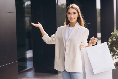 Şık ceketli neşeli bir kadın modern bir mağazanın dışında alışveriş torbaları tutuyor. Davetkar bir şekilde, başarılı bir alışveriş deneyiminin coşkusunu ve heyecanını simgeliyor..