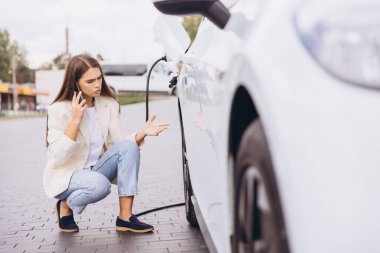 Endişeli bir kadın elektrikli arabasının yanında diz çöker, telefonuyla konuşurken şarj eder. İfadesi, teknoloji ve iletişim temalarını vurgulayarak hüsran ve aciliyet taşıyor..