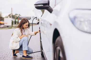 Genç bir kadın elektrikli bir arabanın yanında diz çöküp şarj kablosunu açık hava istasyonuna bağlıyor. Sahnede çevre dostu ulaşım ve günlük yaşam entegrasyonu vurgulanıyor.