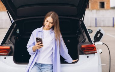 Modern teknolojiyi ve çevre dostu ulaşımı sembolize eden, akıllı bir telefonla elektrikli bir arabaya yaslanmış şık bir genç kadın..