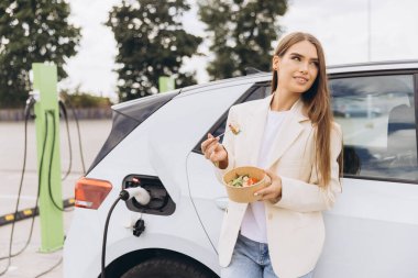 Sağlıklı Salatadan Hoşlanan Genç Kadın, Dışarıdaki Elektrikli Arabaları Şarj Ederken, Çevre dostu Yaşayanlar, Sürdürülebilir Ulaşım ve Şehirdeki Modern Yaşam Tarzı Seçenekleri