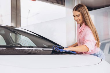 Pembe gömlekli neşeli genç bir kadın araba yıkamada arabasını temizlerken gülümsüyor. Olumlu ifadesi ve odaklanmış ilgisi, aracıyla ilgilenmenin verdiği memnuniyeti vurguluyor..