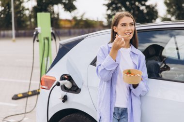 Genç bir kadın açık hava istasyonunda elektrikli arabası şarj edilirken sağlıklı bir salatanın tadını çıkarıyor. Sahne, sürdürülebilirliği, sağlıklı yaşam tarzını ve modern teknolojiyi huzur ortamında yansıtıyor..