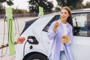 Genç bir kadın açık hava istasyonunda elektrikli arabası şarj edilirken sağlıklı bir salatanın tadını çıkarıyor. Sahne, sürdürülebilirliği, sağlıklı yaşam tarzını ve modern teknolojiyi huzur ortamında yansıtıyor..