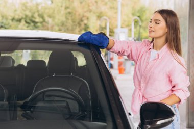 Pembe gömlek giyen genç bir kadın açık hava araba yıkamada arabasını mavi bir bezle temizliyor, gülümsüyor ve işten zevk alıyor. Ayarlar gelişigüzel ve araç bakımına odaklı..