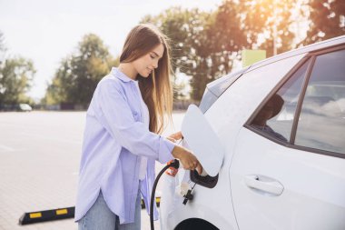 Güneşli bir günde bir otoparkta elektrikli araba şarj eden kadın çevre dostu ulaşım ve sürdürülebilir enerji kullanımı sergiliyor..