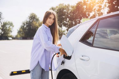 Güneşli bir günde bir otoparkta elektrikli araba şarj eden kadın çevre dostu ulaşım ve sürdürülebilir enerji kullanımı sergiliyor..