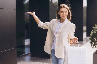 Neşeli bir gülüşü olan şık bir kadın bir şehir binasının önünde el kol hareketi yaparken alışveriş torbaları tutuyor. Güneş ışıl ışıl parlıyor, canlı ve canlı bir atmosfer yaratıyor..