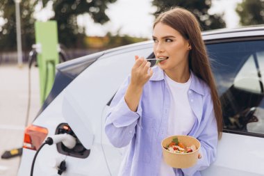Çağdaş şarj istasyonunda elektrikli arabası şarj olurken genç bir kadın taze salatanın tadını çıkarıyor. Bu resim modern yaşam tarzını ve sürdürülebilir yaşamı yakalar..