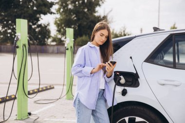 Genç bir kadın şarj istasyonunda akıllı telefonunu kullanarak elektrikli arabasına yaslanıyor. Kurulumda modern teknoloji, sürdürülebilirlik ve kolaylık vurgulanıyor.