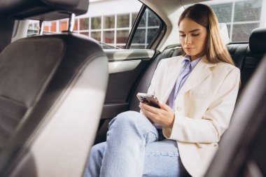 Bej ceketli ve kot pantolonlu neşeli bir kadın arabada otururken akıllı telefonuyla video görüşmesi yapıyor. Keyfi yerinde ve sohbete dalmış görünüyor..