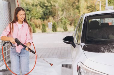 Pembe gömlekli genç bir kadın güneşli bir günde arabasını temizlemek için bir basınç makinesi kullanır. Dış mekan, araba yıkama sürecine tazelik ve canlılık katıyor..