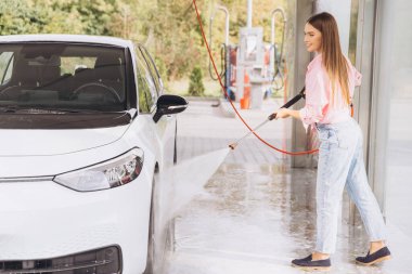 Genç bir kadın yüksek basınçlı hortumla arabasını oto yıkamada zevkle yıkıyor. Aydınlatma güneşli bir günde açık havada, temizliği ve özeni vurguluyor..