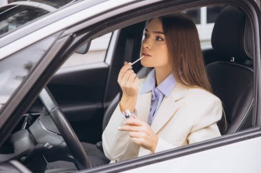 Bir kadın arabasında otururken ruj sürüyor. Açık renkli bir ceket giyiyor ve odaklanmışa benziyor..