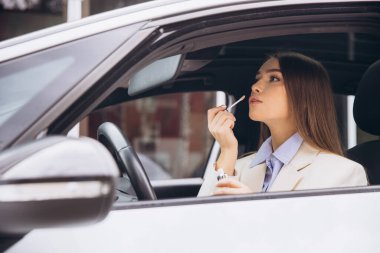 Bir kadın arabasında otururken ruj sürüyor. Açık renkli bir ceket giyiyor ve odaklanmışa benziyor..