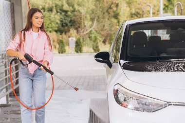 Pembe gömlekli genç bir kadın güneşli bir günde arabasını temizlemek için bir basınç makinesi kullanır. Dış mekan, araba yıkama sürecine tazelik ve canlılık katıyor..