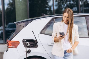 Şık kıyafetli genç bir kadın elektrikli bir arabanın yanında akıllı telefon kullanırken gülümsüyor. Modern teknoloji, çevre dostu ulaşım ve dijital yaşam tarzını temsil ediyor.
