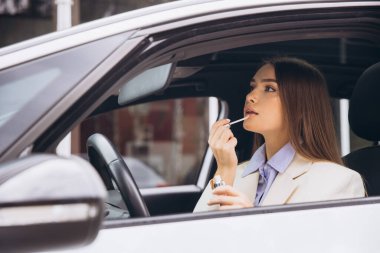 Bir kadın arabasında otururken ruj sürüyor. Açık renkli bir ceket giyiyor ve odaklanmışa benziyor..