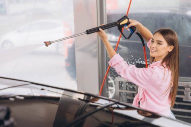 Genç bir kadın self-servis istasyonunda arabasını yıkamaktan hoşlanır. Gülümsüyor, yüksek basınçlı su hortumu kullanıyor ve pembe bir gömlek giyiyor..