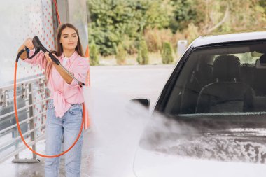 Genç bir kadın oto yıkama servisinde basınçlı yıkama makinesiyle arabasını temizliyor. O, çamaşır yıkama sürecini etkili bir şekilde yönetirken odaklanmış ve memnun görünüyor..