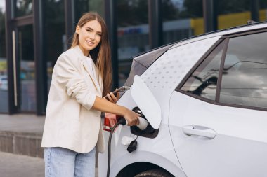 Modern bir istasyonda elektrikli arabasını şarj eden genç bir kadın, güvenle gülümsüyor. Bu resim çevre dostu kavramları, sürdürülebilir enerjiyi ve elektrikli araç teknolojisinin kolaylığını betimliyor..