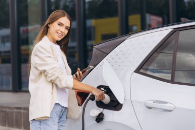 Modern bir istasyonda neşeli genç bir kadın elektrikli arabasını şarj ediyor. Sahne, pozitif ve yenilikçi bir gelecek sunan çevre dostu ulaşım ve sürdürülebilir enerji çözümlerini temsil ediyor.