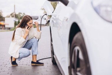 Bir kadın şarj istasyonunda elektrikli arabasının yanında diz çöker ve bir telefon görüşmesi yapar. Modern teknolojiyi ve sürdürülebilir ulaşımı vurgulayarak odaklanmış görünüyor..