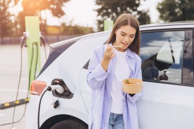 Genç bir kadın, elektrikli arabası istasyonda şarj edilirken sağlıklı bir salatanın tadını çıkarıyor. Öğleden sonra güneş ışığı altında aracın yanında rahatça duruyor..