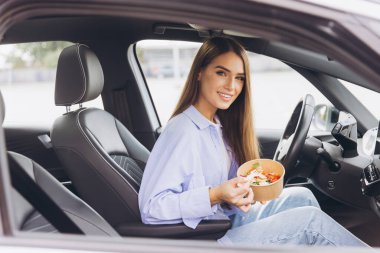 Genç bir kadın arabasında oturmuş sağlıklı bir salata yiyor, yoğun bir günde hızlı ve besleyici bir yemeğin özünü yakalıyor..
