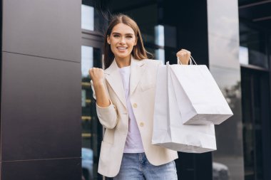 Beyaz ceketli neşeli bir kadın alışveriş torbaları tutuyor, modern bir şehir ortamında heyecan ve başarıyı ifade ediyor. Perakende terapi ve tüketici memnuniyeti kavramları için mükemmel..