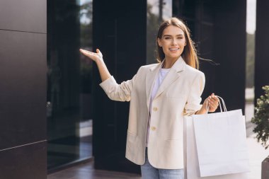 Neşeli bir kadın gündelik ceket giyiyor, alışveriş torbalarını modern bir mağazanın önünde tutuyor. Sahne, alışverişten haz ve heyecan yayıyor. Perakende ve moda kavramları için mükemmel..
