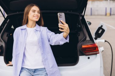 Neşeli genç bir kadın elektrikli arabasının yanında selfie çekiyor, modern bir otoparkta şarj oluyor. Günlük giysiler giyiyor, eko-arkadaşlığı ve çağdaş yaşam tarzını yansıtıyor..