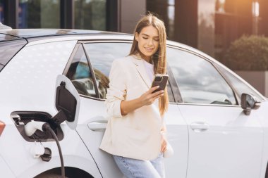 Genç bir kadın akıllı telefonuyla elektrikli arabasının yanında duruyor ve modern bir yaşam tarzının tadını çıkarıyor. Şehir ortamında aracını şarj ederken memnun görünüyor. Çevre dostu ve teknolojiye odaklı tema.