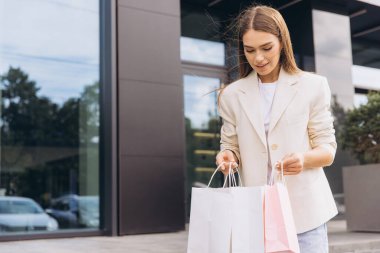 Genç bir kadın, modern bir binanın dışında birkaç alışveriş çantası tutarken gülümsüyor. Başarılı bir alışveriş gezisinden sonra tatmin olmuş ve tatmin olmuş görünüyor..