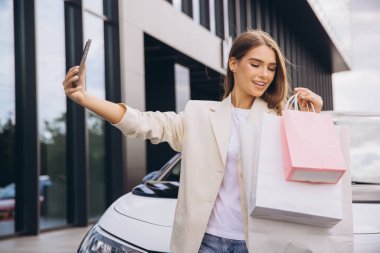 Modern bir binanın önünde elinde alışveriş torbalarıyla selfie çeken neşeli bir kadın. Mutluluk ve başarı yayıyor, tatmin edici bir alışveriş gününü kutluyor..