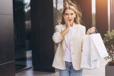Bir mağazanın önünde elinde alışveriş torbalarıyla neşeli bir kadın duruyor. Şaşkınlık ve mutluluk ifadesi modern bir şehir ortamında alışveriş yapmanın heyecanını yansıtıyor..