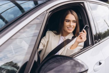 Kendine güvenen genç bir kadın lüks arabasında oturuyor, elinde arabanın anahtarları var ve gülümsüyor. Keyifli bir yolculuğun tadını çıkarırken özgürlük ve bağımsızlık yayıyor. Sürüş ve başarı kavramları için mükemmel görüntü.