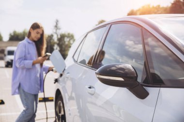 Mavi gömlekli genç bir kadın, güneşli bir otoparkta elektrikli bir arabayı şarj ederken telefonunu kullanıyor, çevre dostu ulaşımı vurguluyor..