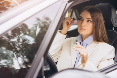 Arabadaki profesyonel bir kadın rimel sürüp özgüven ve çoklu görev becerilerini somutlaştırır. İş elbisesi giyiyor, iş günü için hazırlanıyor. Ayarlar, yoğun ve modern bir yaşam tarzı olduğunu gösteriyor..