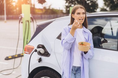 Genç bir kadın, elektrikli arabası istasyonda şarj edilirken sağlıklı bir salatanın tadını çıkarıyor. Öğleden sonra güneş ışığı altında aracın yanında rahatça duruyor..