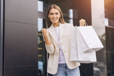 Günlük kıyafetli genç bir kadın başarılı alışveriş gezisini yumruk pompasıyla kutluyor, alışveriş torbalarını mağazanın önünde tutuyor, neşe ve memnuniyeti ifade ediyor..